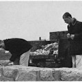 Two young men with rocks gathered in a cart next to a stone wall.