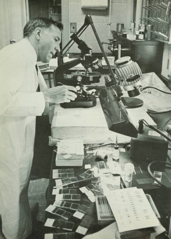 A man in a white lab coat works at a microscope.