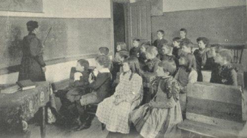 A teacher points at the blackboard in front of a class of children.