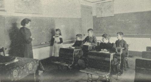 A teacher and a girl stand at a blackboard.  Four boys sit at their desks.