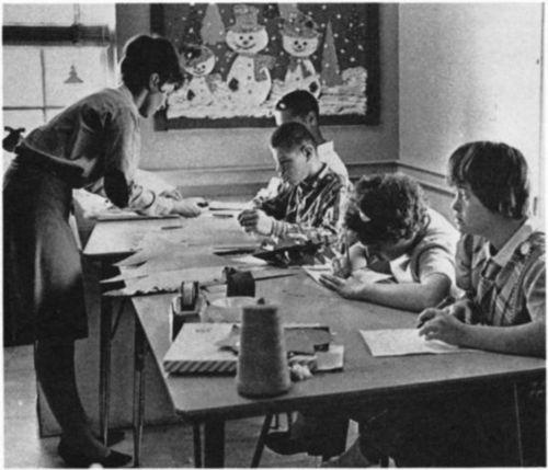 A woman helping four children working on art.