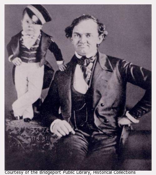 P.T Barnum sits in a chair.  Tom Thumb, with hand on Barnum's shoulder and in uniform, stands on table.