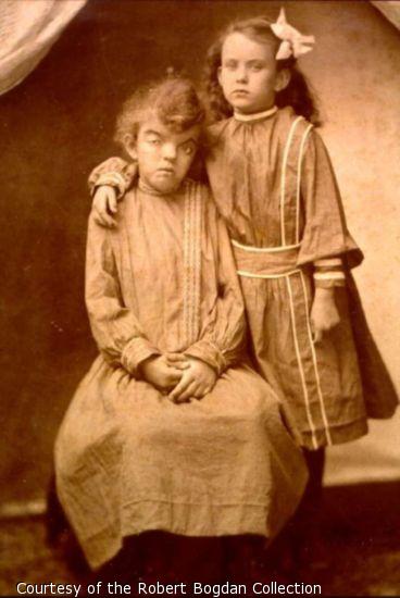 One sister sits on a stool with her hands folded in her lap. The other stands with her arm around her sister.