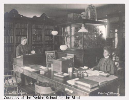 Two women at desks, one sitting, one standing.