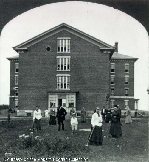 Stereo card showing male and female patients, including children, playing crocquet in front of a large brick building at Willard Asylum.