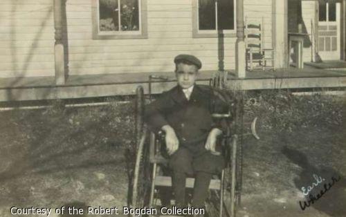 Photograph of a boy using a wheelchair in the yard of a small house.