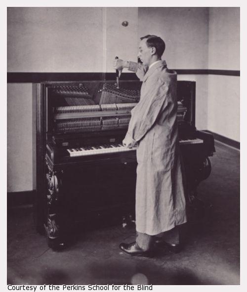 Man in white lab coat stands in profile at piano, working with tuning instruments.