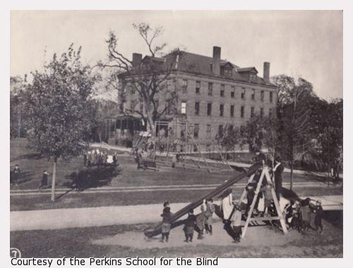 Children playing on a playground with slide, with Perkins Institution for the Blind in background.