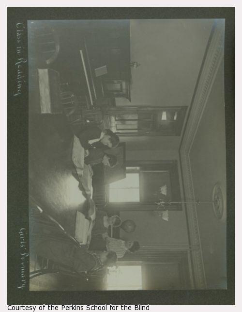 Four girls in dark dresses reading at large table, teacher in light dress standing.