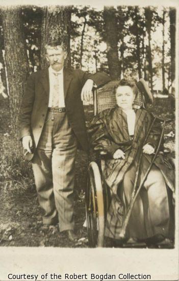 Woman sitting in wheelchair and holding crutch. Man stands next her holding cigar.