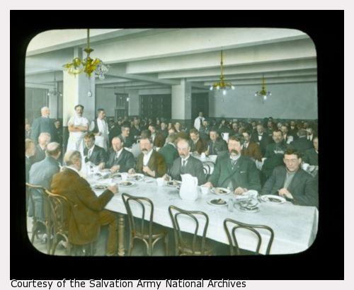 Large dining hall, men serving food to men with suit jackets at long tables, tablecloths set.