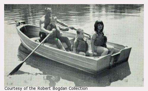 Photograph of young man helping a boy row a rowboat.  Two girls are also in the boat.