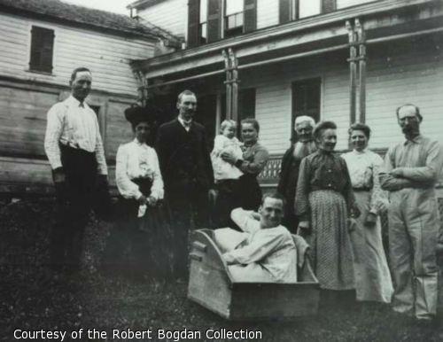 Photograph of a family posing outside of a house.  A young man wearing a night shirt sits in a wooden box.
