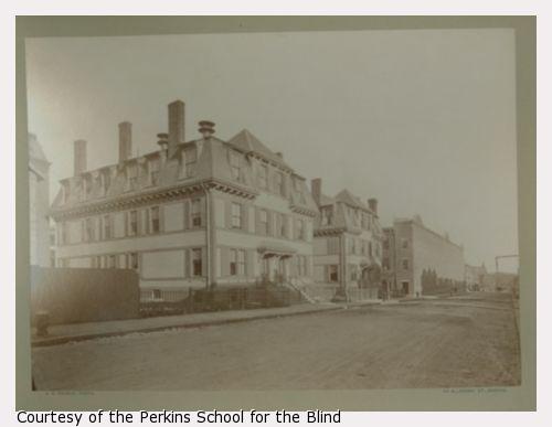 Row of cottages used as residential quarters at the Perkins Institution and Massachusetts School for the Blind in South Boston. There are two, side-by-side, three story cottage buildings that face a wide unpaved road.