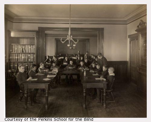 Classroom filled with boys at the Perkins Institution and Massachusetts School for the Blind in South Boston. They are seated around a large U-shaped wooden table with an assortment of plants, pine cones, and flowers. Each boy has a writing tablet and writing instrument.