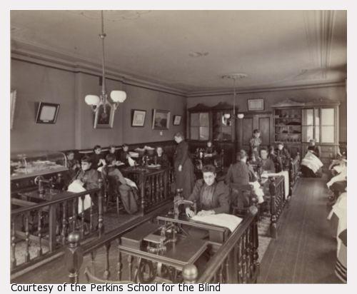 Girl's sewing class from upper grammar at the the Perkins Institution and Massachusetts School for the Blind in South Boston. The sewing room has eight or more sewing machine tables in two rows with railings sectioning off the work stations. Students sit on benches along the walls with sewing in their laps.