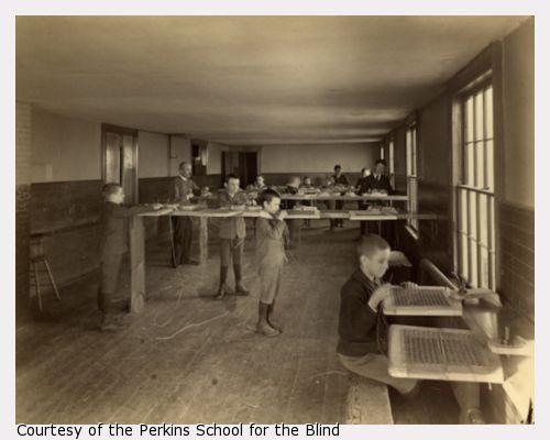 Boys' caning shop, Main building, Perkins Institution, South Boston, Mass. Showing Mr. Thomas J. Carroll, instructor. The boys stand at tall work benches with chair seats to be caned clamped at chest height. They each have long lengths of caning materials that they weave across the wooden frames.