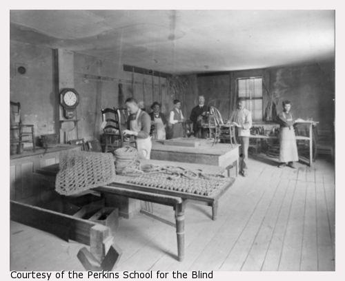 Workshop for the Blind, Perkins Institution for the Blind, Fourth Street, South Boston, Mass. In front are two thick door mats and a spool of the thick cord used to make them. Six men stand in the back of the room at work stations. Some work on chair caning. One of the faces has been enhanced with pen for publication purposes.