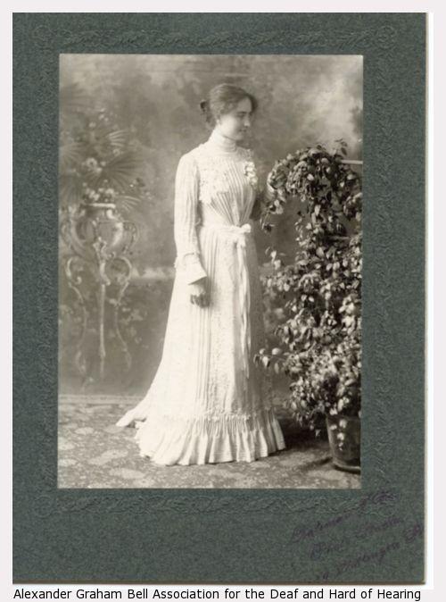 Helen Keller standing, facing right, wearing long sleeve, floor length, white gown with waist sash, facing plant.