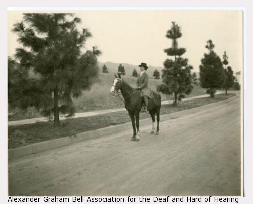 Helen Keller riding horseback on a pine tree-lined road.