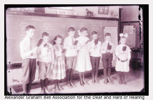 Both male and female deaf children stand in front of a chalk board to read at the Horace Mann School.
