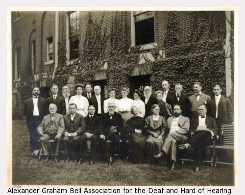 American Association To Promote Teaching Speech To The Deaf 1906 Convention photograph taken outdoors. One row of attendees sit and one row of attendees stands in front of a large wall with climbing ivy.