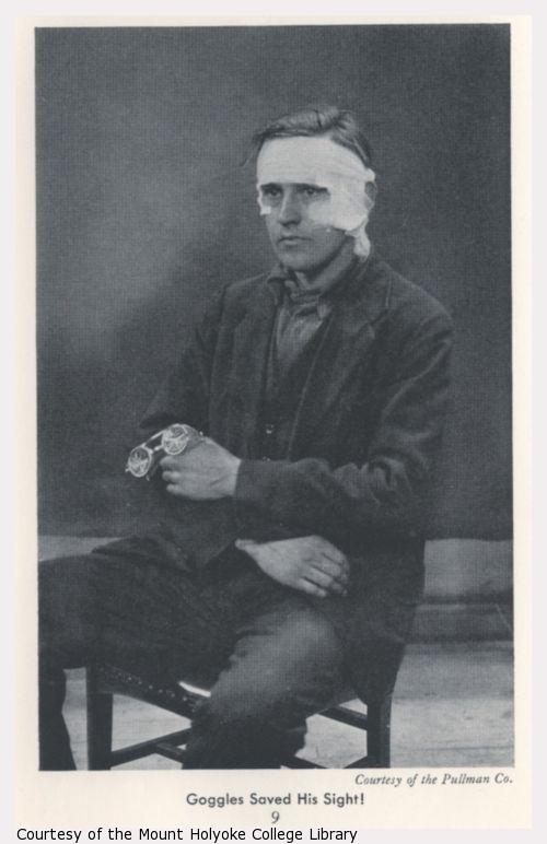 Man sitting in chair, face bandaged, holding broken pair of safety goggles.