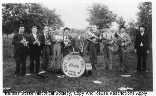 A nine-piece brass band stands in a field in front of trees holding their instruments.  They stand around a large drum on which is printed "State Insane Asylum Band, Topeka, KS".