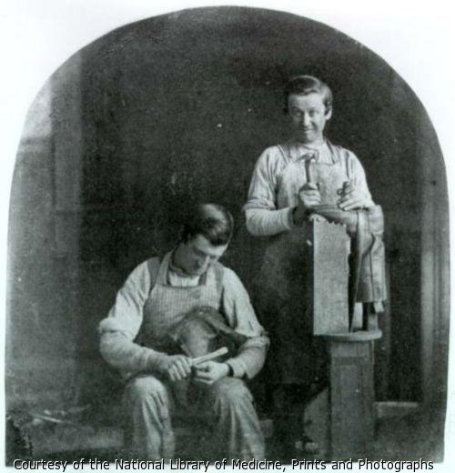 Two boys working as cobblers. One files the sole of a shoe. The other holds a hammer and last upright and smiles.