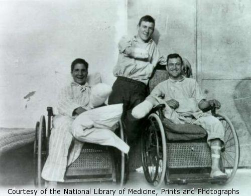 Three smiling veterans. Two leg amputees are seated in wicker wheelchairs. An arm amputee stands behind them.