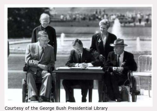 George Bush in front of White House sitting at table signing ADA; with a priest, a woman, and two men in wheel chairs
