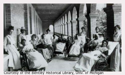 Seven women sit in large wheelchairs on the porch of a large Victorian house at a sanitarium. Each is covered in a blanket and looks passively at the camera. Six attendants and two children stand behind and amongst them.
