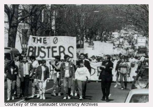 A crowd of protesters marches down a street.  A man holds a banner that reads "The Board Busters."
