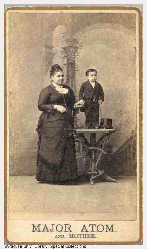 Major Atom, holding a top hat, stands on a table next to an average sized woman.