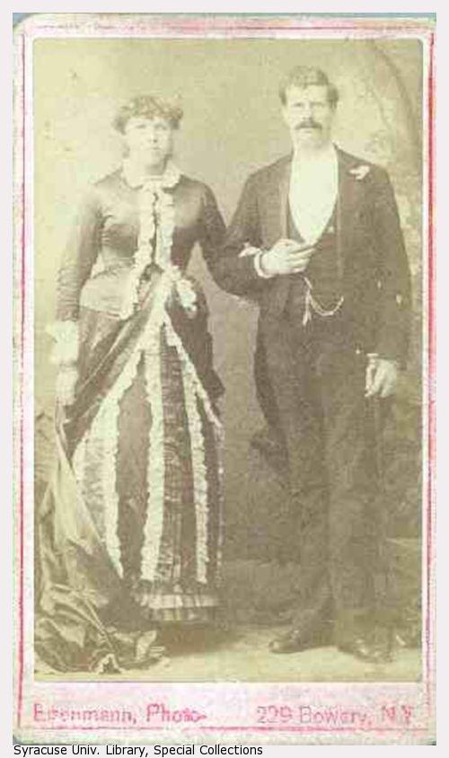 A portrait of a couple standing, the woman in a dress with ruffles and the man in a suit.
