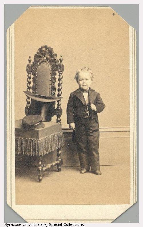 A short-statured young man in formal attire stands next to a chair.