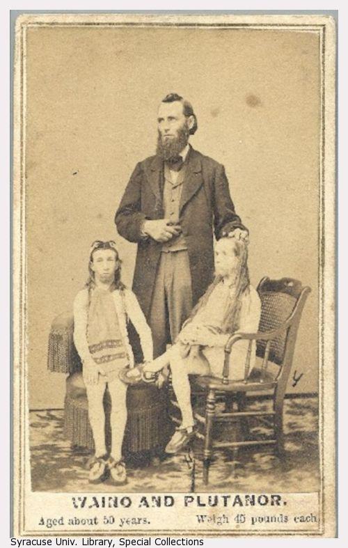 An average-sized man stands between two sitting short-stautured men with long hair and beards.
