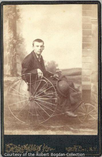 A young man, with hat on his knee, sits in a wheelchair with metal wheels.