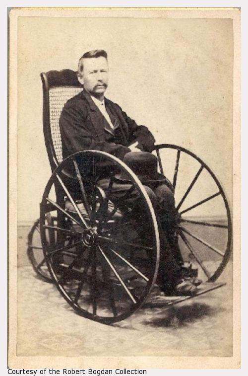 A man, with a hat on his lap, sits in a wheelchair.